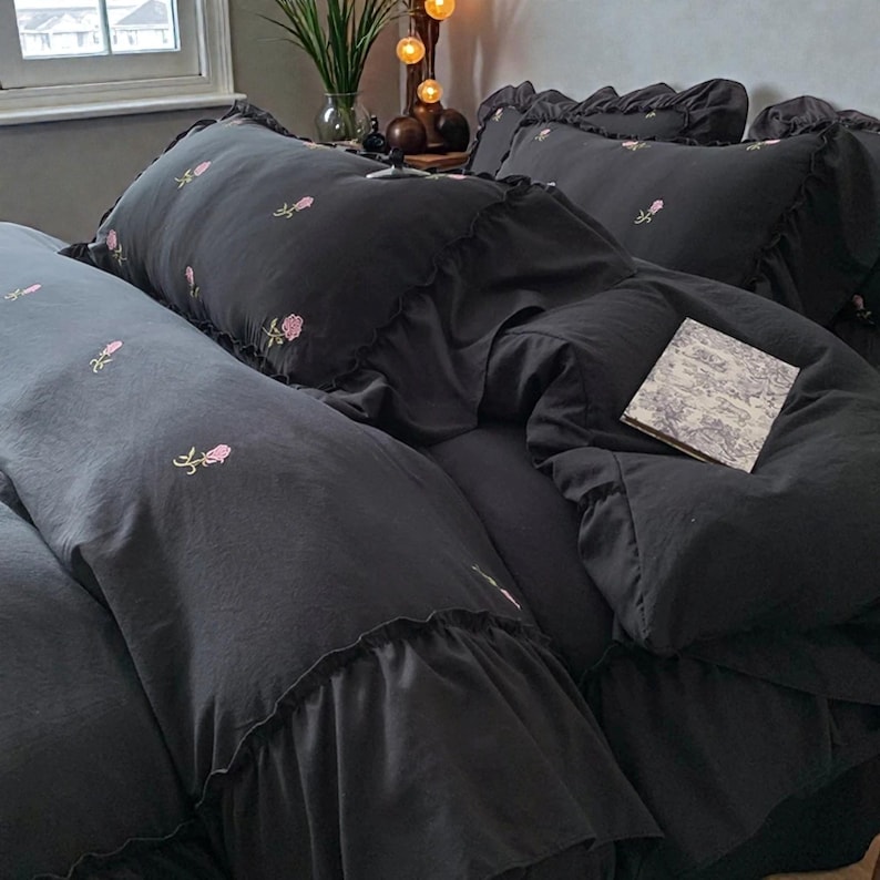May include: Dark gray bedding set with embroidered pink roses. The set includes a comforter, pillows, and a long bolster pillow. The comforter and pillowcases have ruffled edges. A small book with a floral design rests on the comforter.