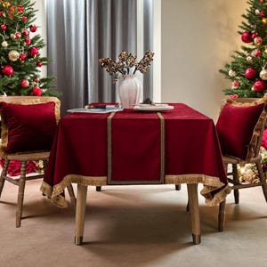 Può includere: Una scena di sala da pranzo festosa con una tovaglia rossa e cuscini abbinati su sedie di legno. Il tavolo è apparecchiato con un vaso bianco, piatti e un runner decorativo. Alberi di Natale con ornamenti rossi e dorati fiancheggiano il tavolo, creando un'atmosfera calda.