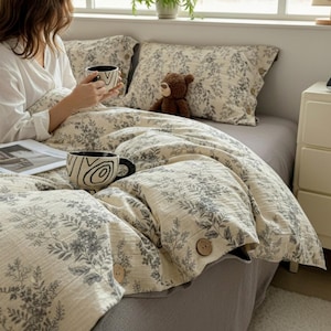 Può includere: Set di biancheria da letto beige e grigio a fantasia floreale, composto da due federe e un copripiumino. Il copripiumino ha bottoni in legno. Un orsacchiotto marrone e due tazze bianche e nere sono sul letto.
