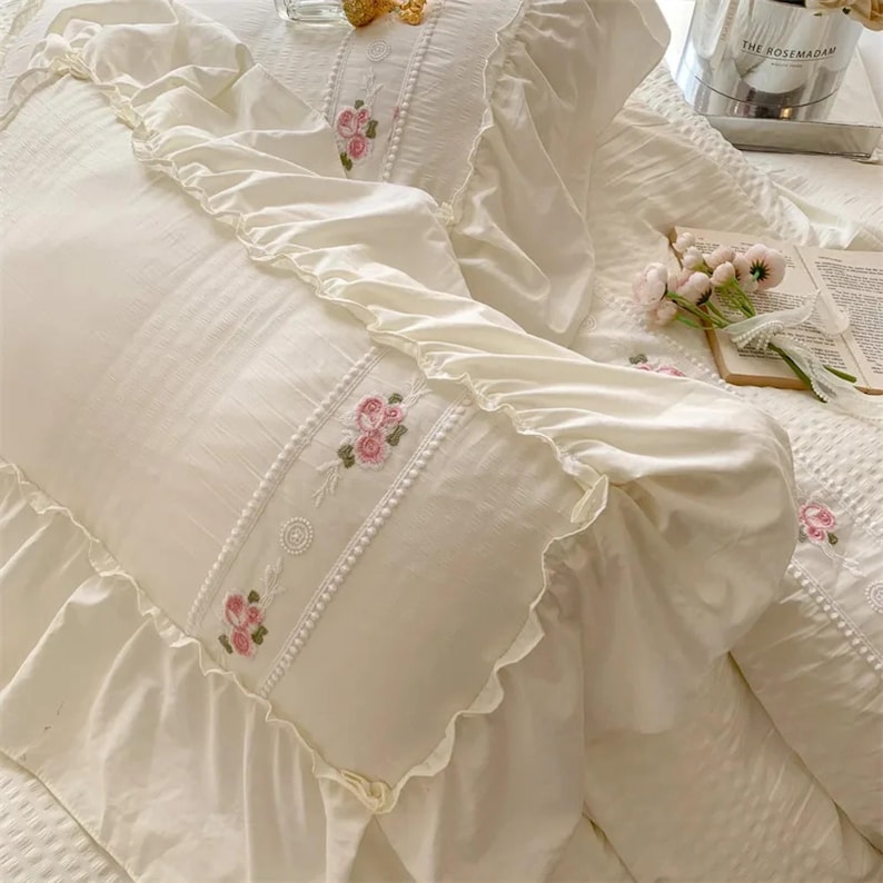 May include: Ivory-colored bedding set featuring embroidered pink roses. The set includes a ruffled pillowcase and a duvet cover with a textured pattern. The pillowcase has a decorative embroidered band with a pearl-like trim.