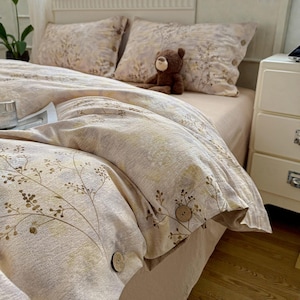Può includere: Set di biancheria da letto beige e marrone chiaro con motivo floreale, con copripiumino e federe abbinate. Il copripiumino presenta grandi bottoni in legno. Un piccolo orsacchiotto marrone è seduto sul letto.