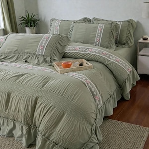 May include: Sage green bedspread and pillow set with ruffled edges. The bedding features a vertical ribbed pattern and a decorative floral trim in white and pink. A wooden tray with a grapefruit sits on the bed.
