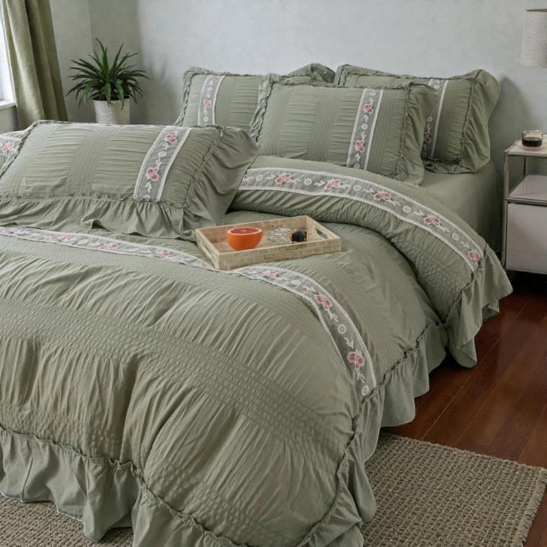 May include: Sage green bedspread and pillow set with ruffled edges. The bedding features a vertical ribbed pattern and a decorative floral trim in white and pink. A wooden tray with a grapefruit sits on the bed.