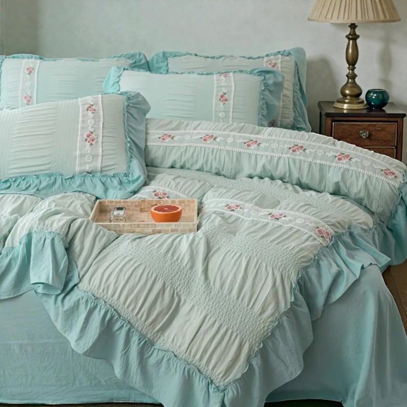 May include: A light blue bed set with ruffled edges and floral embroidery. The set includes pillows, a comforter, and a bed skirt. A wooden tray with a grapefruit sits on the comforter. The bedding has a vintage, cottagecore aesthetic.