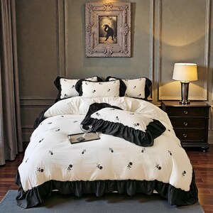 May include: A white and black floral bedding set with a duvet cover, pillowcases, and decorative pillows. The duvet and pillowcases have a black ruffle trim. The bedding set is on a bed in a bedroom.