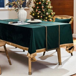 May include: A dark green velvet tablecloth with gold fringe trim, set on a wooden table. The table is set for a festive occasion, with a Christmas tree in the background. A white vase with branches is also on the table.