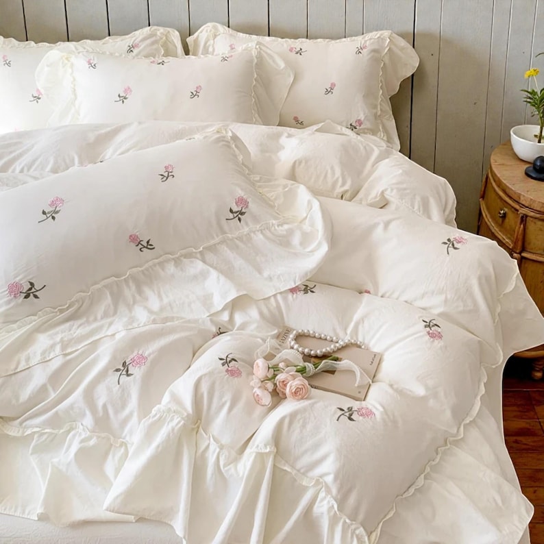 May include: White bedding set with embroidered pink floral accents. The set includes a duvet cover, pillows, and a ruffled edge. A pearl necklace and a small bouquet of pink roses rest on the bed, creating a soft, romantic aesthetic.