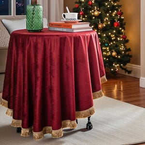 Op de afbeelding: Een ronde, bordeauxrode fluwelen tafelkleed met gouden franjes bedekt een kleine tafel. Een groene glazen fles, boeken en een kopje thee staan erop. Een kerstboom met versieringen staat op de achtergrond.