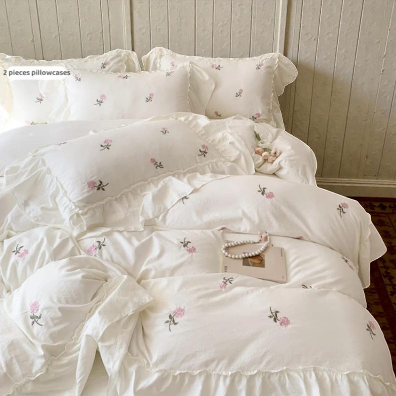 May include: White bedding set with embroidered pink floral accents. The set includes two pillowcases, a duvet cover, and additional pillows. The bedding has a ruffled edge and a delicate, romantic aesthetic. The text "2 pieces pillowcases" is visible.