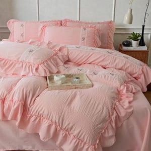 May include: A pink bedding set with ruffled edges and embroidered details. The set includes a comforter, pillows, and a bed skirt. A decorative tray with a candle and small items sits on the bed. The overall theme is romantic and feminine.