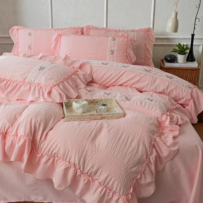May include: A pink bedding set with ruffled edges and embroidered details. The set includes a comforter, pillows, and a bed skirt. A decorative tray with a candle and small items sits on the bed. The overall theme is romantic and feminine.