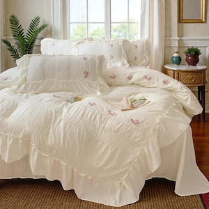 May include: Cream-colored bedding set with ruffled edges and delicate pink floral embroidery. The set includes a comforter, pillowcases, and decorative pillows. The bed is made up with a skirted base, creating a soft and inviting aesthetic.