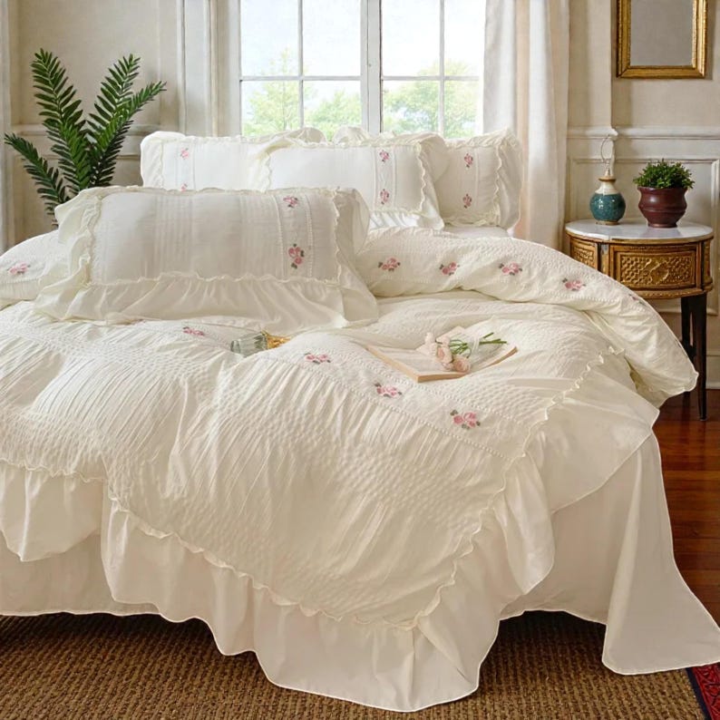 May include: Cream-colored bedding set with ruffled edges and delicate pink floral embroidery. The set includes a comforter, pillowcases, and decorative pillows. The bed is made up with a skirted base, creating a soft and inviting aesthetic.