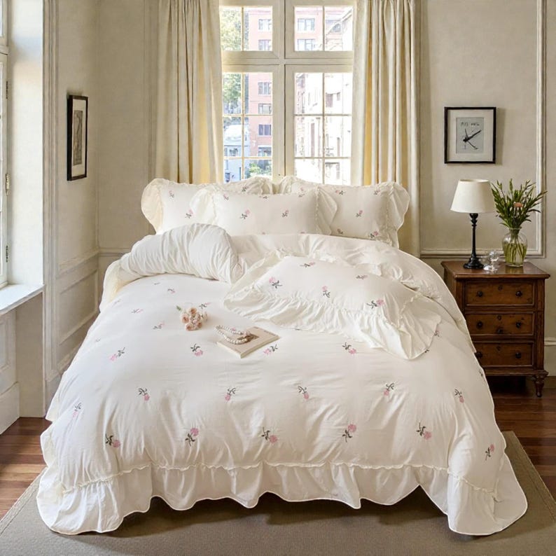 May include: A white bed set with pink floral embroidery. The set includes a comforter, pillows, and a ruffled bed skirt. The bedding is arranged on a bed in a well-lit room with a window and curtains.