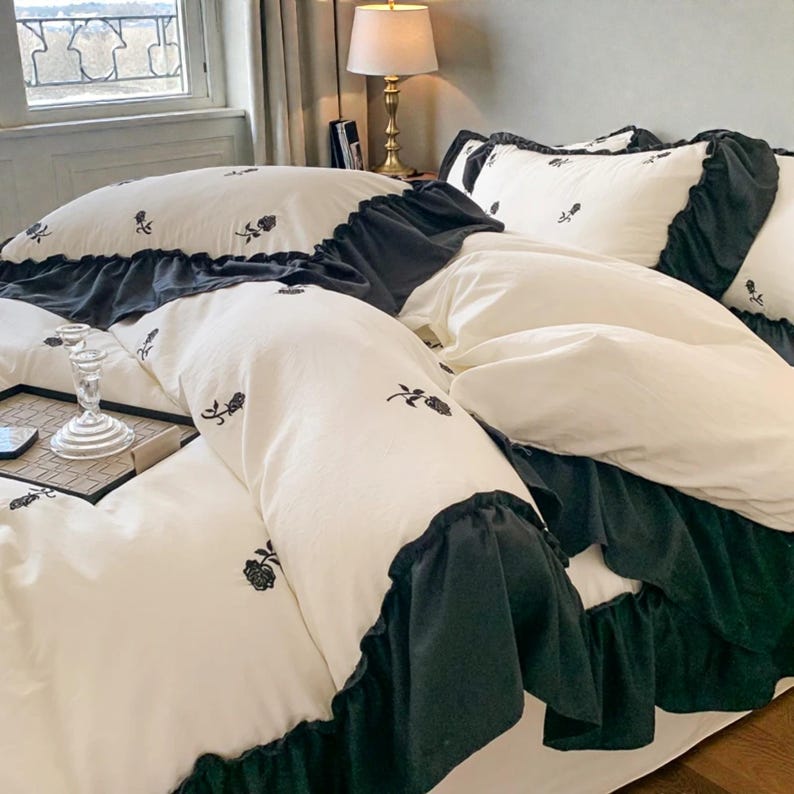 May include: White bedding set with black floral embroidery and a dark gray ruffled trim. The set includes a duvet cover and pillowcases. The bedding is made of a soft, comfortable material, perfect for a bedroom.