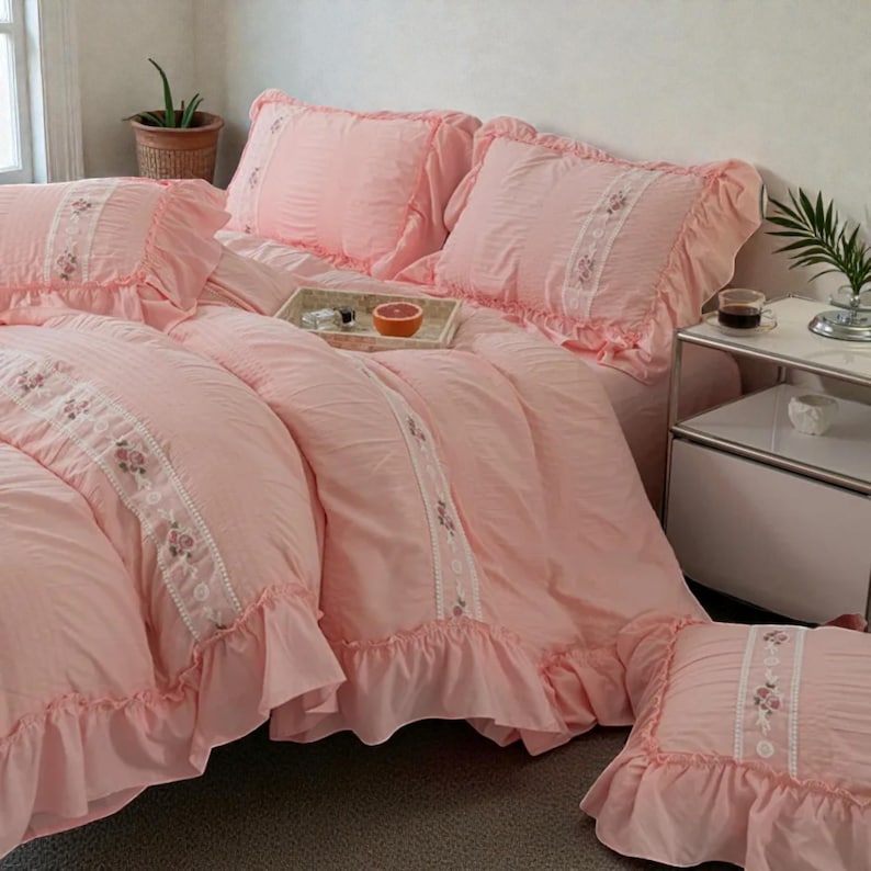 May include: A pink bedding set with ruffled edges and floral embroidery. The set includes a comforter, pillows, and a decorative pillow. A tray with a grapefruit half and a cup of coffee sits on the bed. The room has a soft, inviting atmosphere.