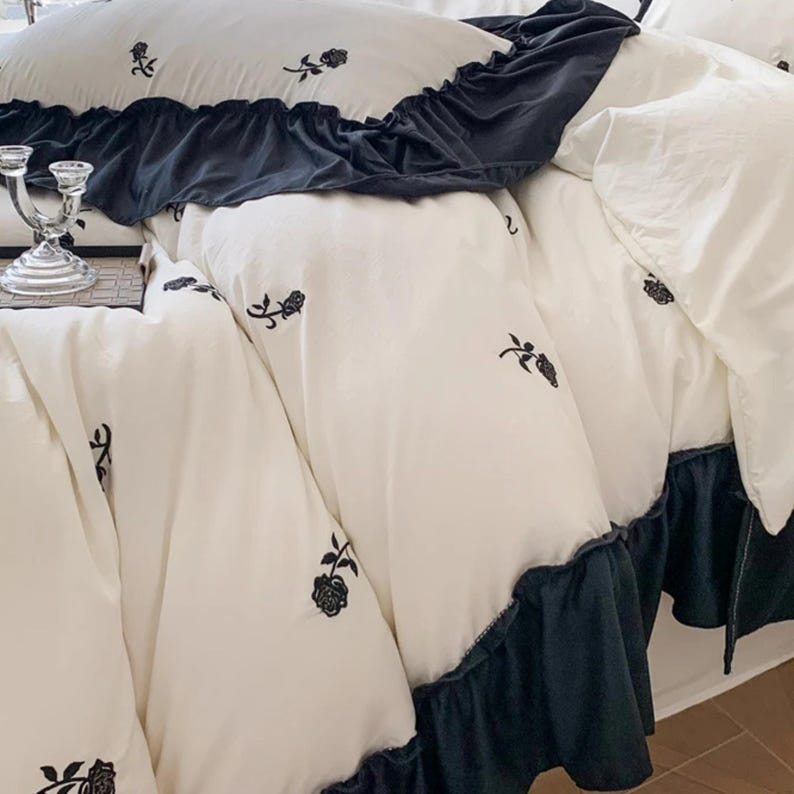 May include: White bedding set with black floral embroidery. The set includes a duvet cover, pillowcases, and a bed skirt with a dark blue ruffle. The floral pattern is repeated across the pieces, creating a cohesive and elegant look.