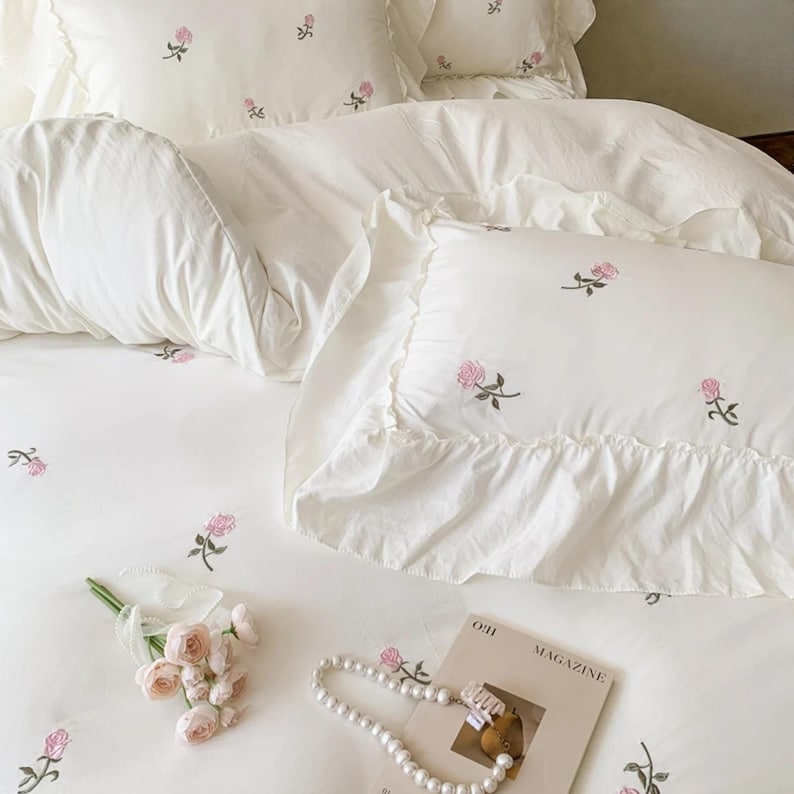 May include: White bedding set with embroidered pink roses. The set includes a duvet cover and ruffled pillowcases. A bouquet of pink roses, a pearl necklace, and a magazine are arranged on the bed, creating a romantic and elegant aesthetic.