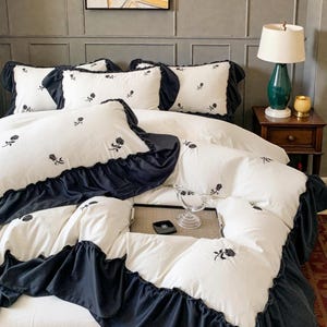 May include: White bedding set with black ruffled trim and embroidered rose details. The set includes several pillows and a duvet cover. The bed is made up with a white sheet and a dark blue bed skirt. The room has a lamp and a side table.