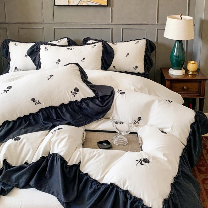 May include: White bedding set with black ruffled trim and embroidered rose details. The set includes several pillows and a duvet cover. The bed is made up with a white sheet and a dark blue bed skirt. The room has a lamp and a side table.