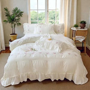 May include: A white bed set with ruffled edges and floral embroidery. The bedding includes a comforter, pillows, and a duvet cover. The room has a window with natural light, a potted plant, and a small side table.