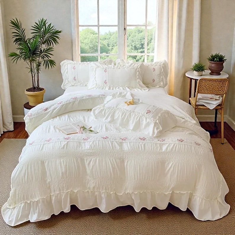 May include: A white bed set with ruffled edges and floral embroidery. The bedding includes a comforter, pillows, and a duvet cover. The room has a window with natural light, a potted plant, and a small side table.