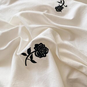May include: Off-white fabric with a subtle texture, featuring embroidered black roses. The roses are detailed with petals and leaves, scattered across the surface. The fabric is draped, creating soft folds and shadows.