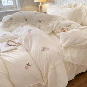 May include: Cream-colored bedding set featuring embroidered pink roses. The set includes a duvet cover, pillowcases, and a decorative pillow, all with ruffled edges. A pearl necklace and a book are placed on the bed, enhancing the romantic aesthetic.