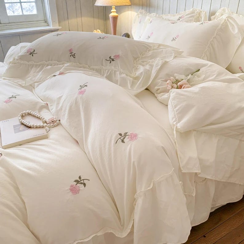 May include: Cream-colored bedding set featuring embroidered pink roses. The set includes a duvet cover, pillowcases, and a decorative pillow, all with ruffled edges. A pearl necklace and a book are placed on the bed, enhancing the romantic aesthetic.