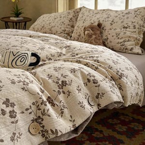 Può includere: Set di biancheria da letto beige a fantasia floreale con copripiumino, federe e cuscino decorativo. Il copripiumino ha grandi bottoni in legno. Una tazza decorativa con manico nero è sul letto. Un piccolo peluche è sui cuscini.