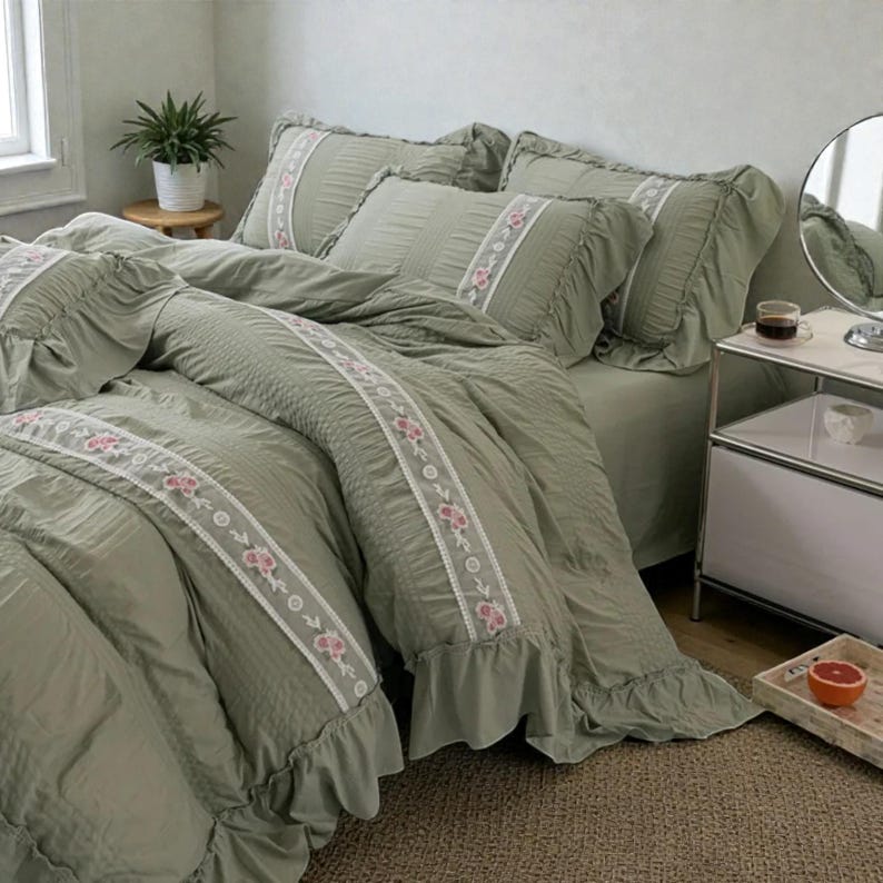 May include: Sage green bedding set with ruffled edges and embroidered floral details. The set includes a comforter, pillowcases, and a bed skirt. The comforter and pillowcases feature a white and pink floral embroidered stripe.