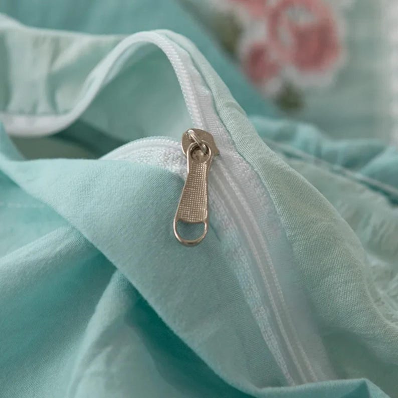 May include: Close-up of a light blue fabric with a white zipper. The zipper pull is silver. Faint floral patterns in pink and green are visible in the background. The fabric appears to be soft and possibly part of bedding.
