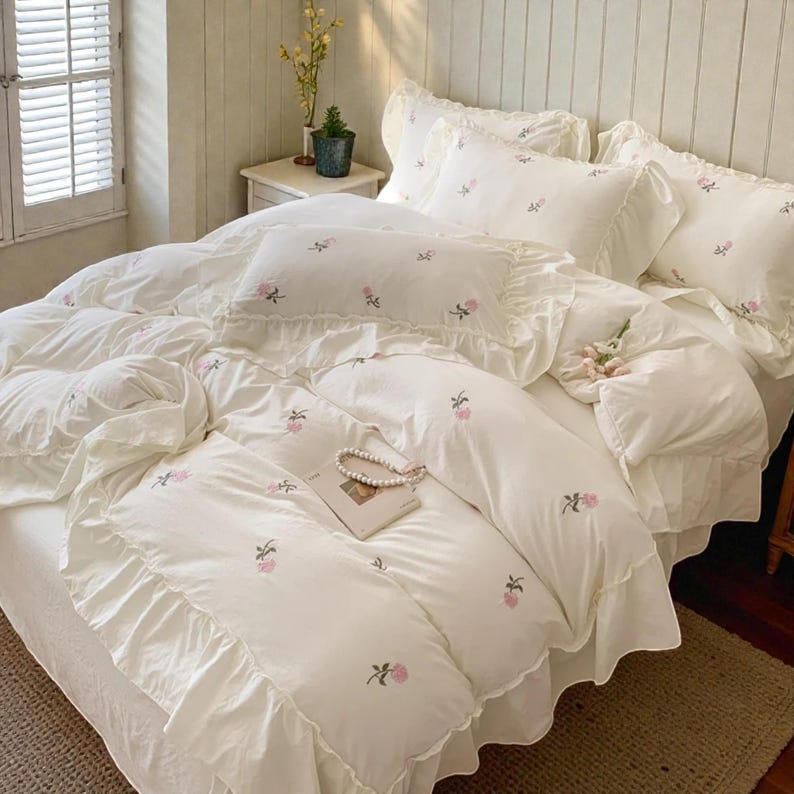 May include: A bed is covered with a white floral embroidered bedding set. The set includes a duvet cover, pillowcases, and a bed skirt, all adorned with delicate pink rose embroidery and ruffled edges. A book and a pearl necklace rest on the bed.