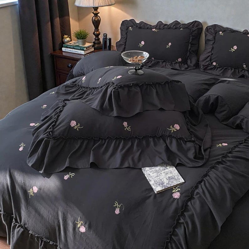 May include: Dark gray bedding set with embroidered pink roses. The set includes a comforter, pillows, and a decorative pillow with ruffled edges. A small book rests on the comforter. The bedding is arranged on a bed.