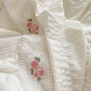 May include: Close-up of white fabric with embroidered pink flowers and green leaves. The fabric has a textured, striped pattern on the right side and a floral embroidered border on the left side. The overall theme is delicate and feminine.