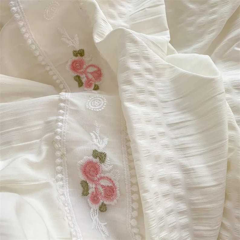 May include: Close-up of white fabric with embroidered pink flowers and green leaves. The fabric has a textured, striped pattern on the right side and a floral embroidered border on the left side. The overall theme is delicate and feminine.