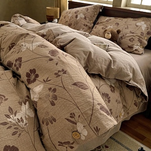 Peut inclure: Un ensemble de literie avec un motif floral beige et marron. La housse de couette et les taies d'oreiller présentent un motif floral répété. Un petit ours en peluche est posé sur les oreillers. La housse de couette a des boutons en bois.