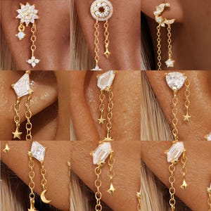 Puede incluir: Una colección de pendientes dorados con varios diseños celestiales y geométricos. Algunos pendientes presentan cadenas colgantes con estrellas, lunas y detalles de cristal. Los pendientes se muestran en una oreja, destacando sus delicados detalles.