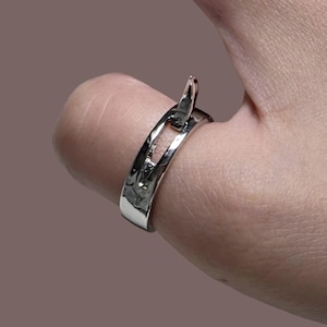 Puede incluir: Un anillo plateado con un diseño único, con un elemento elevado en forma de gancho. El anillo tiene una textura martillada y se lleva en un dedo. El fondo es marrón liso y apagado.