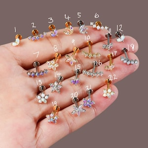 May include: A selection of ear piercing studs in various designs, shown on a hand. The studs have gold and silver finishes, with floral, crescent, and geometric shapes, and are adorned with colourful gemstones.