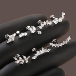 May include: A selection of silver ear piercing jewellery. The earrings showcase diverse designs, including leaf, teardrop, and geometric shapes, all embellished with clear, sparkling gemstones. The jewellery is presented against a dark backdrop.