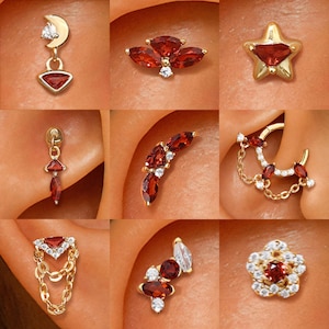 Pode incluir: Uma coleção de brincos dourados com vários designs, incluindo uma lua crescente, uma estrela e formas florais. Cada brinco apresenta pedras preciosas vermelhas e brancas, mostrando uma variedade de estilos para piercings na orelha.