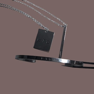 Könnte beinhalten: Eine silberfarbene Halskette mit einem rechteckigen Anhänger und einem passenden Armband. Der Anhänger und das Armband tragen beide die Buchstaben "BTS". Der Schmuck wird vor einem braunen Hintergrund präsentiert.