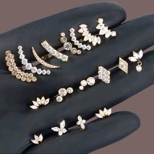 Può includere: Una collezione di gioielli per il corpo color oro, tra cui orecchini a cerchio, bilancieri e borchie. I gioielli sono adornati con cristalli trasparenti e scintillanti in varie forme, come cerchi, foglie e farfalle. I pezzi sono esposti su una superficie scura.