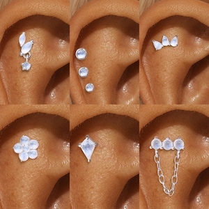 Puede incluir: Una colección de pendientes de plata con varios diseños, incluyendo un grupo de tres piedras redondas, una flor, una forma de diamante y un diseño de cadena. Los pendientes están engastados con piedras iridiscentes, mostrando una gama de estilos.