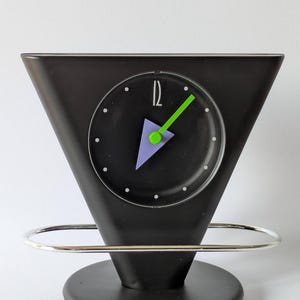 Puede incluir: Una taza de cerámica blanca con asa negra y el texto "But First, Coffee" en negrita negra. La taza se encuentra sobre un fondo borroso, sugiriendo un ambiente acogedor.