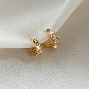 May include: A pair of gold-coloured hoop earrings. The earrings feature a bamboo-like design with textured details. The earrings are presented on a white, textured surface.