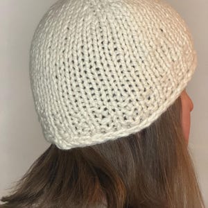 Könnte beinhalten: Eine cremefarbene Mütze aus grobem Strick. Die Kopfbedeckung zeichnet sich durch ein schlichtes, abgerundetes Design mit einem durchgehend sichtbaren glatten Rechtsstrickmuster aus, das diesem Winteraccessoire ein dickes und gleichmäßiges Aussehen verleiht.