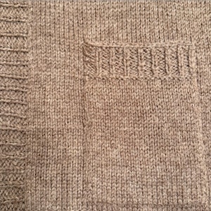 Può includere: Primo piano di un capo in maglia di colore beige neutro. L'immagine mostra la trama del tessuto a maglia, con un bordo a coste a sinistra e una piccola tasca a destra. La maglieria appare morbida e calda.