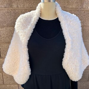 May include: A cream-colored faux fur shrug with a thick, textured, and plush pile. The garment features a wide collar and open front design, draped over a solid black sleeveless top on a mannequin, showcasing a soft and voluminous appearance.